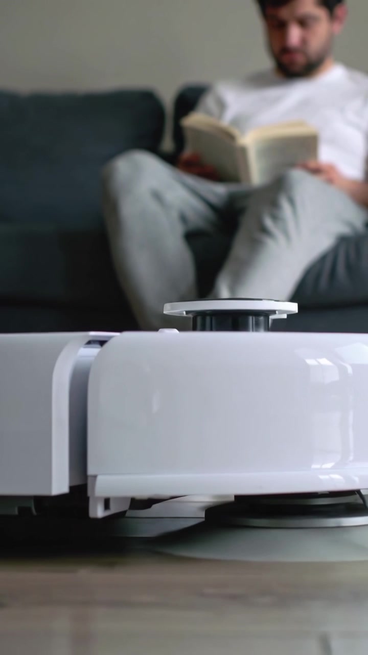 Vertical video: Starting robot cleaning hardwood floor near sofa, reader in white tee reading book