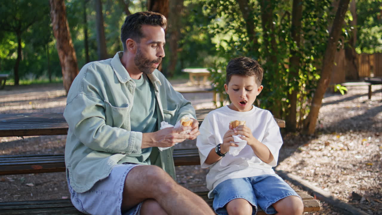 Happy family eating sandwiches on sunny recreational zone. Boy chewing food
