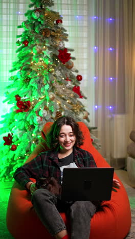relaxed caucasian woman sitting in armchair pets a cat and watches film. relaxing time spending at christmas evening. vertical video.