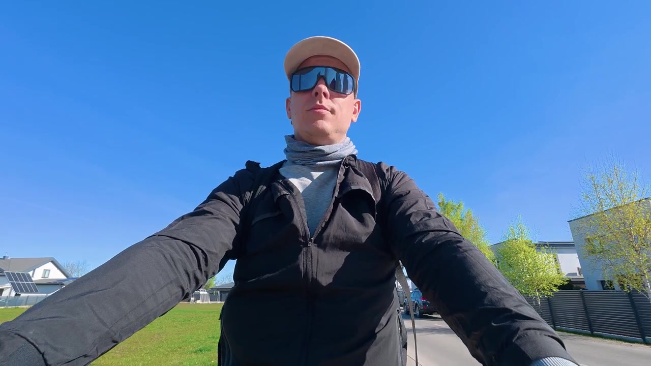 Punto di vista frontale di un uomo in bicicletta attraverso un quartiere suburbano tranquillo e moderno in una giornata di sole. Luce del giorno limpida, cielo azzurro chiaro e case residenziali creano un'atmosfera esterna rilassata e positiva.