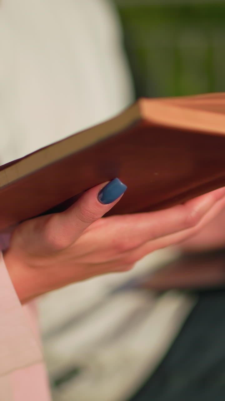 primer plano de una mujer de color rosa leyendo un libro al aire libre, manos manicuradas visibles, amiga borrosa escribiendo en la computadora portátil, fondo verde borroso que mejora el enfoque