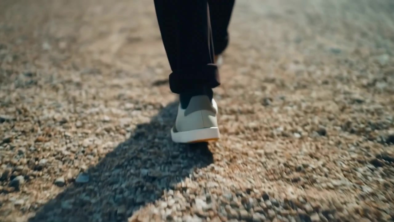 Close view follows lower legs and shoes moving steadily forward on small stones, with warm directional light, shallow focus, and elongated shadows emphasizing each step.