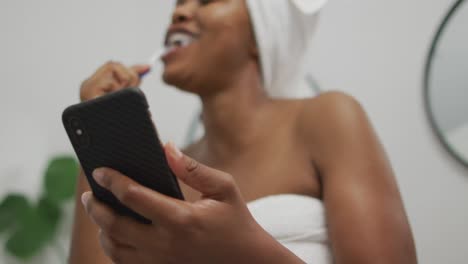 feliz mujer afroamericana cepillándose los dientes y usando un teléfono inteligente en el baño