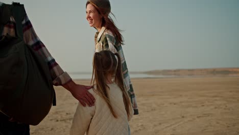 Rear-view-of-a-happy-family,-a-middle-aged-man-in-a-checkered-shirt-walks-along-the-seashore-with-his-wife,-a-brunette-girl,-and-a-little-blonde-daughter-in-a-white-jacket-outside-the-city-during-their-picnic-and-hike-in-the-summer