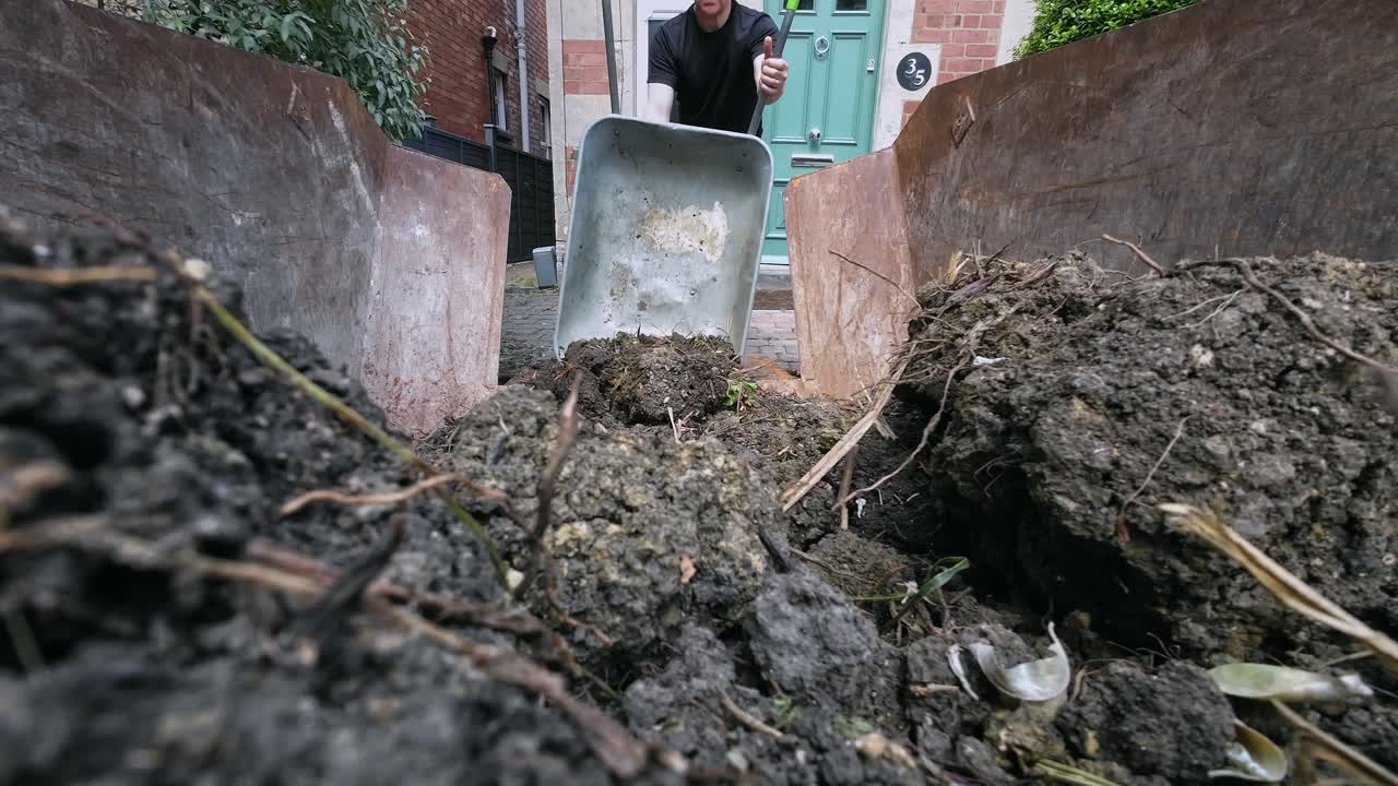 Wide shot of fit, middle aged man wheeling and emptying a wheelbarrow full of soil in to a skip, then using a spade to clear up
