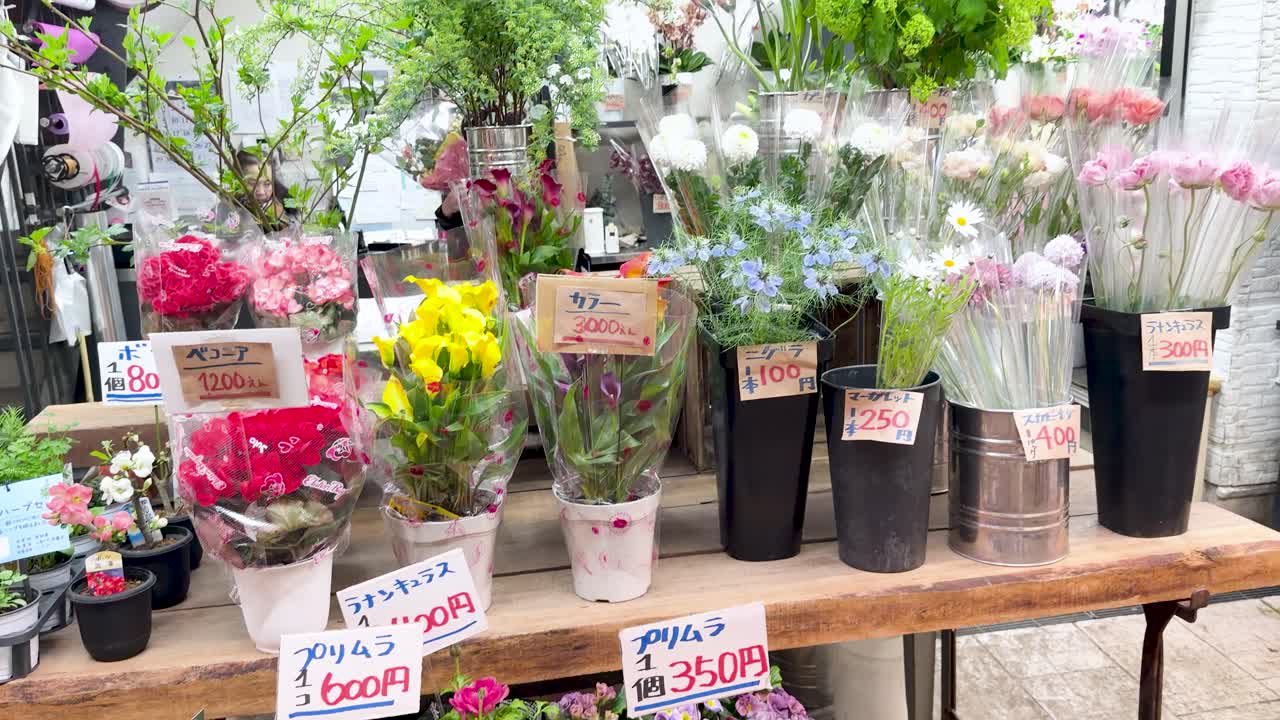 屋外の日本の花屋で、鮮やかな生け花や鉢植えの間を手持ちカメラがパンする。