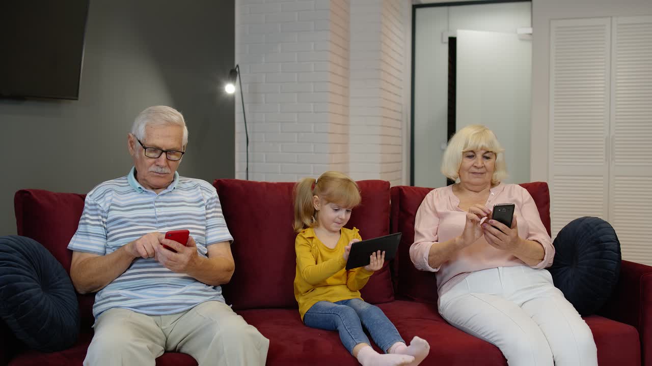 Grandparents and grandchildren sit on the couch at home and use mobile phones and digital tablet