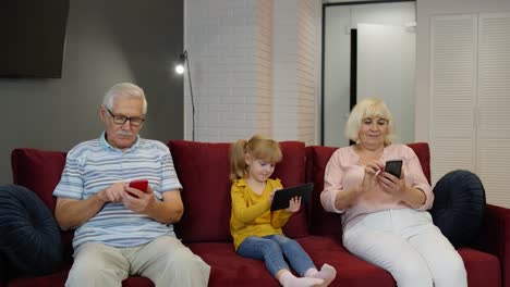 los abuelos y nietos se sientan en el sofá en casa y usan teléfonos móviles y tabletas digitales