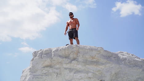 shirtless bodybuilder with sunglasses and cap on the hill in a sunny day. strong athlete with muscular body under blue sky outdoors.