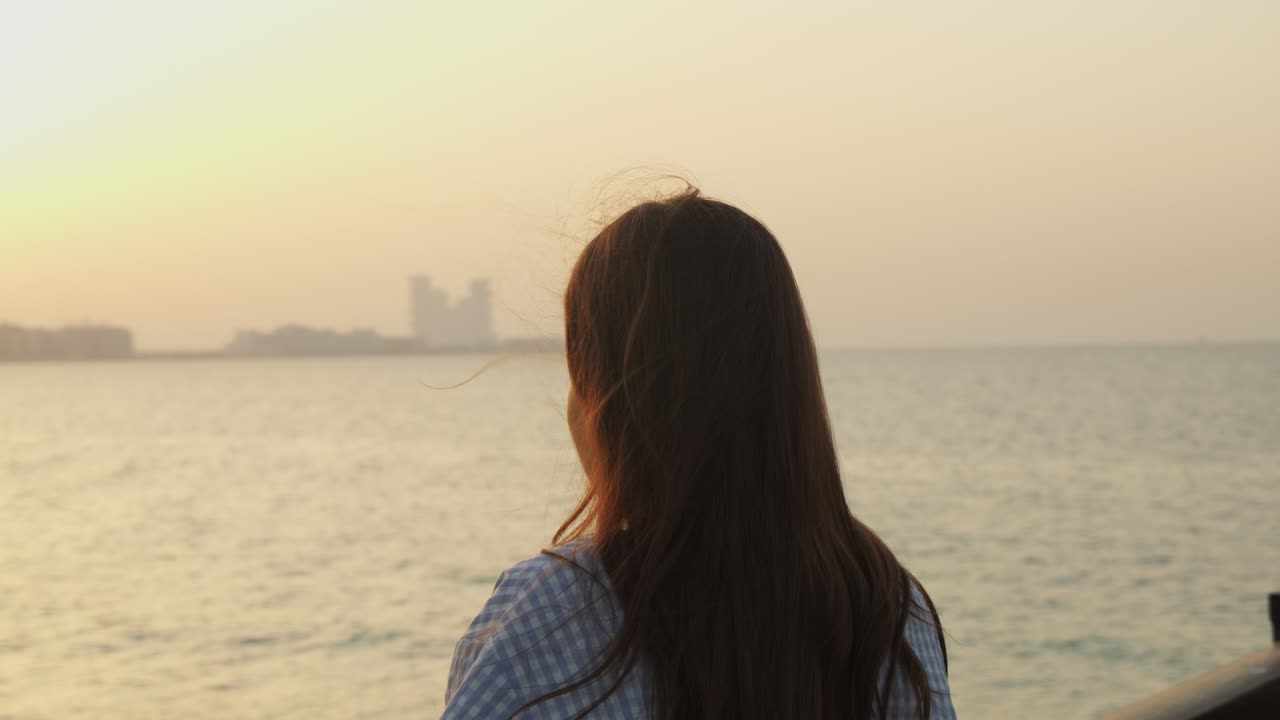 Attractive woman looking at city skyline across the ocean. Orbiting woman with long brown hair looking at Dubai skyline during sunset