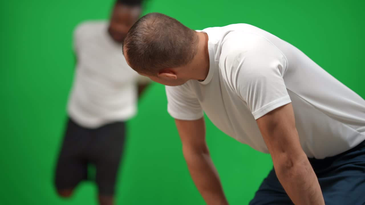 Tired Caucasian sportsman resting as blurred African American man warming up at background. Multiethnic sports team of football players at chromakey green screen background.