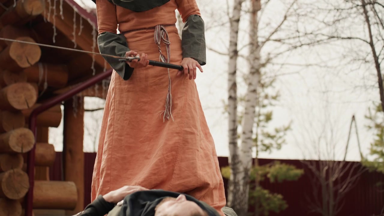 Ung kvinna i medeltida läder- och tygdräkt ligger på mage på torrt gräs bredvid en trähydda medan en stående följeslagare med svärd och lång klänning förblir synlig i en dramatisk historisk utomhusmiljö.