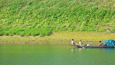 bengali fishermen and villagers rowing a long pirogue along a slowly flowing river in front of a vividly vegetalised riverbank, bangladesh