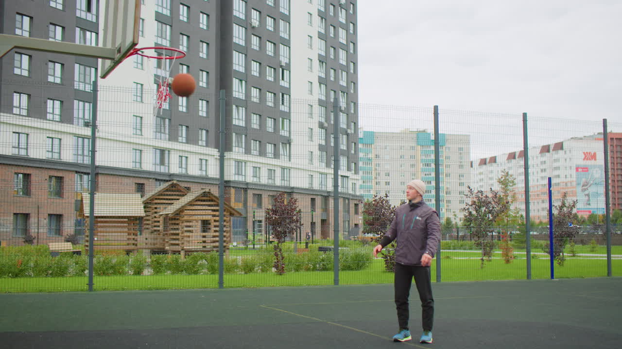 caucasian senior man shooting free throws on urban court, wearing jacket and sneakers, solitary practice under cloudy sky, apartment buildings and metal fence in background, ball hits rim and bounces
