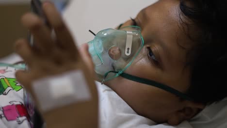 Children-patients-on-bed-with-oxygen-mask-in-hospital-while-playing-cell-phone