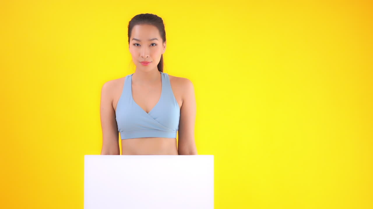 Pretty asian woman in fitness outfit showing empty white board to camera, isolated on yellow background, slow motion
