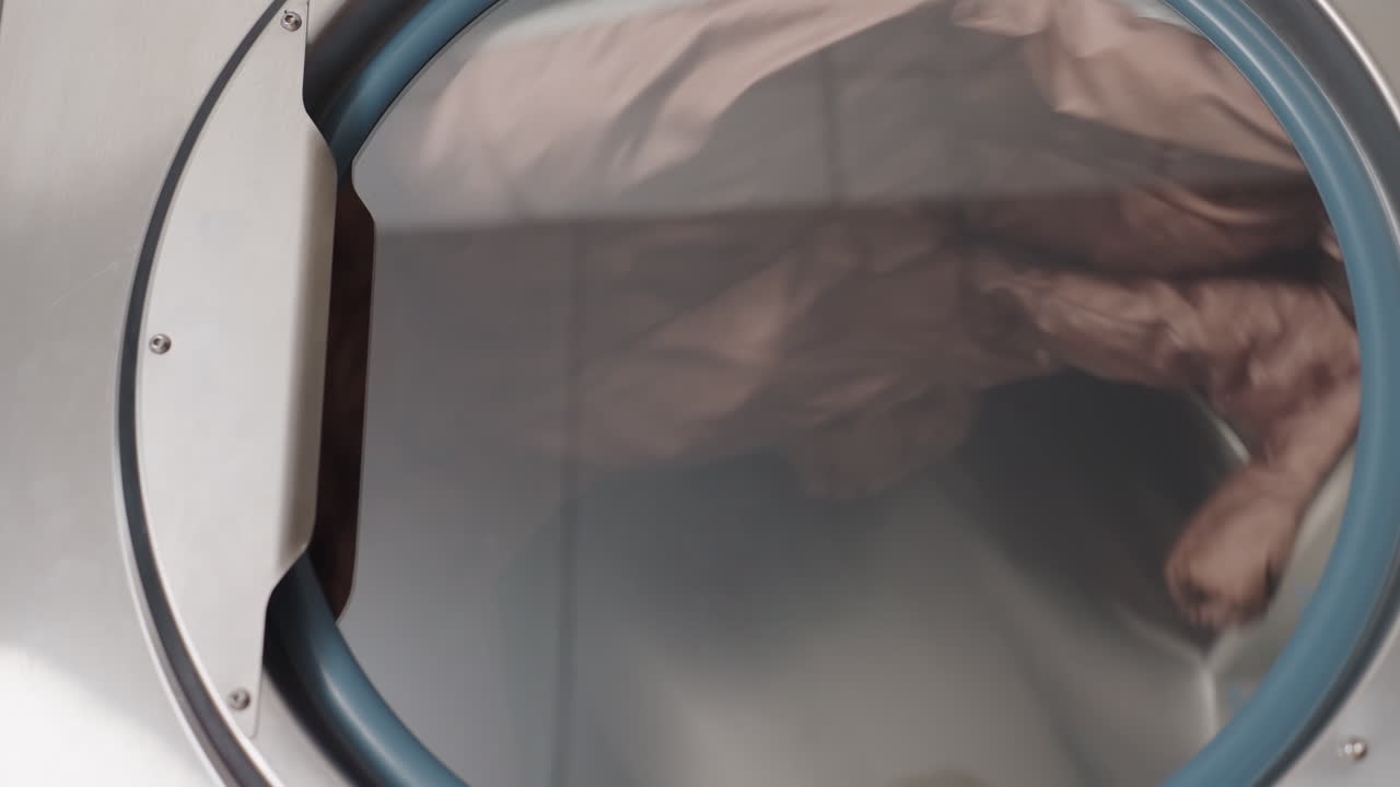 Industrial dryer labeled 13 with round door and blue gasket, inner view shows cloth tumbling during dry cycle, stainless drum spinning inside laundromat, moisture evaporating, airflow through glass