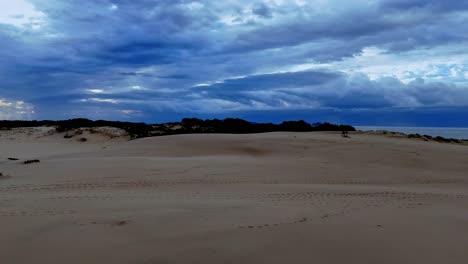 Una-Playa-De-Arena-Con-Algunas-Nubes-En-El-Cielo-Y-Algunas-Plantas-Creciendo-En-La-Arena