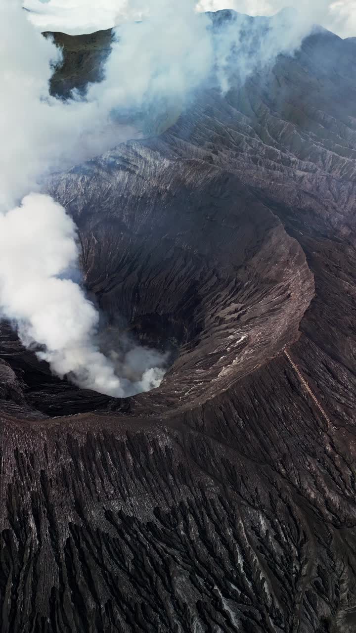 Vertical drone video of Mount Bromo crater showing steep volcanic slopes, dark ash terrain and white steam rising from the crater, filmed from above highlighting active volcano landscape in East Java