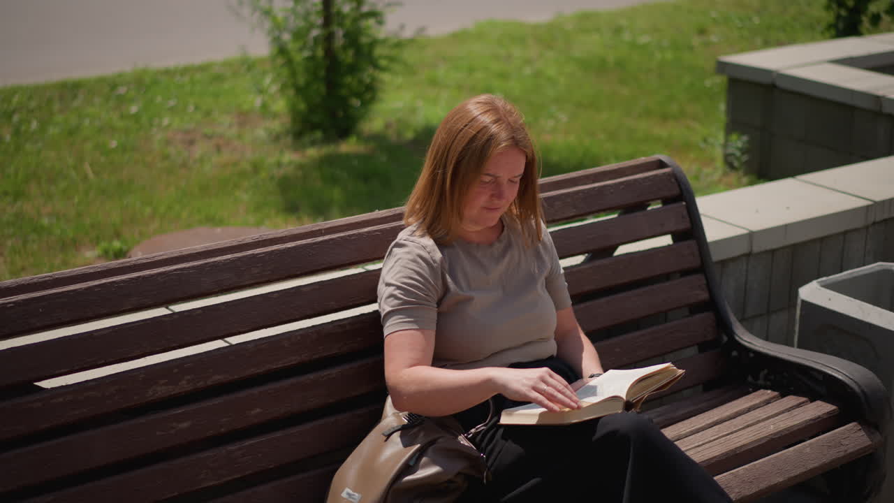Ein Student sitzt auf einer Holzbank und blättert im Freien unter warmem Sonnenlicht in einem aufgeschlagenen Buch, mit einem konzentrierten Gesichtsausdruck und in ruhiger Stimmung, umgeben von Grün