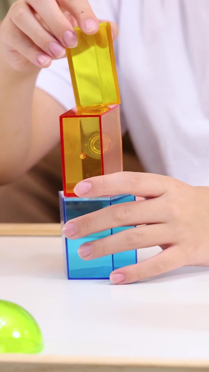 A child's hands carefully stack colorful translucent geometric shapes on a white table surface.
