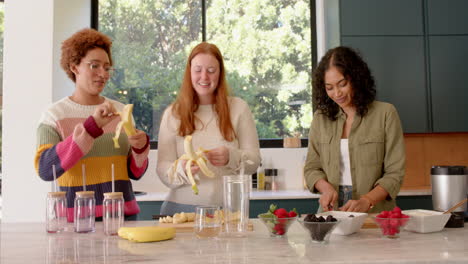 preparing smoothie, diverse female friends peeling bananas and chopping fruits in kitchen