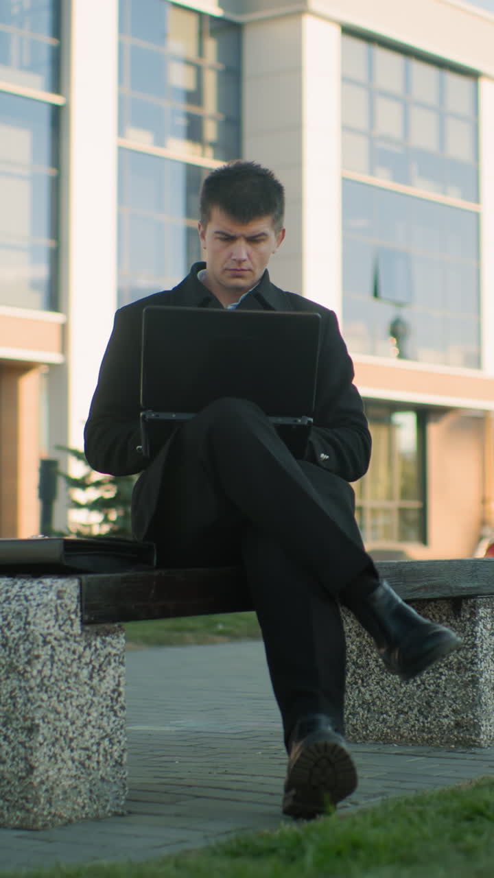 현대적인 오피스 빌딩 앞에 다리를 고 노트북으로 작업하는 야외 벤치에 앉아있는 사업가. 도시 배경에는 초록색, 차량 및 깨한 건축 반사물이 포함됩니다.