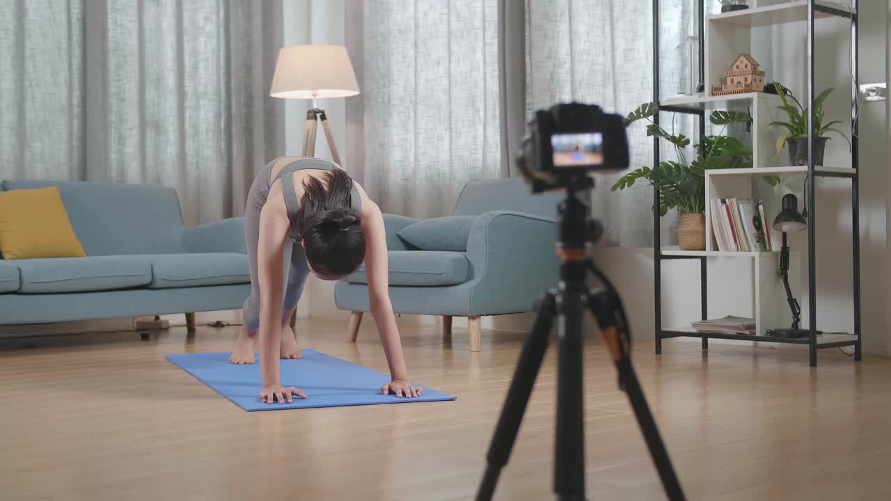 vista completa de joven entrenadora asiática mujer en ropa deportiva hablando a la cámara y estirándose mientras graba ejercicio de enseñanza en casa