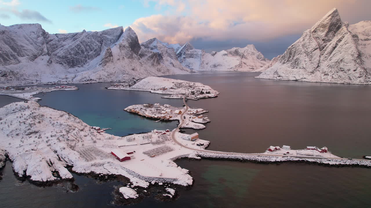 Cinematic 4K drone shot pulling away diagonally from Sakrisoy fishing village. High angle reveal of yellow rorbuer cabins, E10 highway, and snow capped Reinefjorden peaks at Arctic sunset