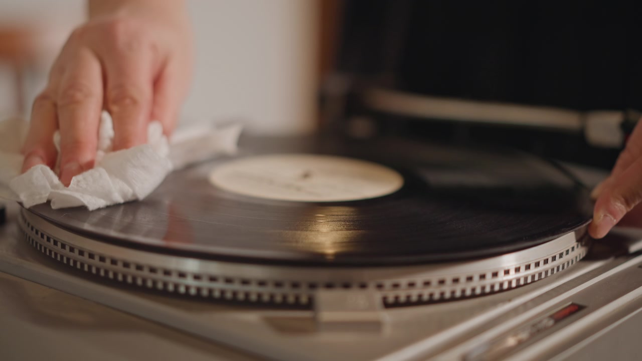 Pulizia ravvicinata di un disco in vinile su giradischi, mani che puliscono delicatamente il disco con un panno morbido sotto una calda luce ambientale in un accogliente salotto. Un meticoloso rituale di restauro da parte di un collezionista, con attenzione ai solchi e alla trama.