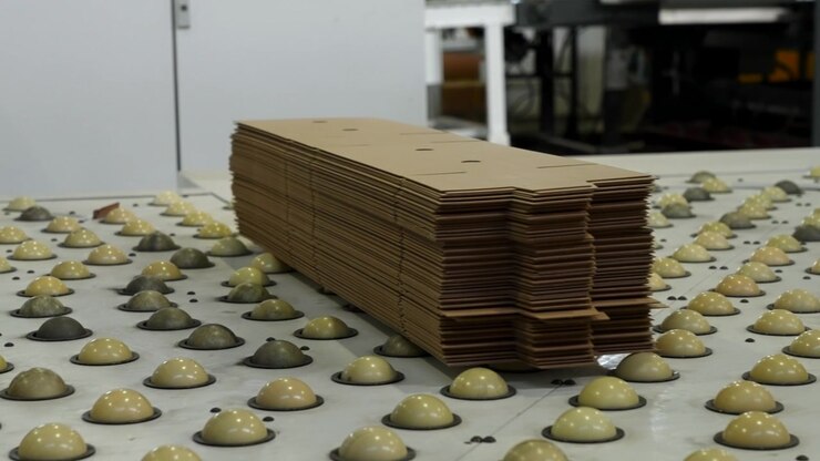 Cardboard sheets are stacked on workshop machine table while dome shaped molded components sit in neat rows nearby, shown in centered perspective with muted tones and soft indoor lighting