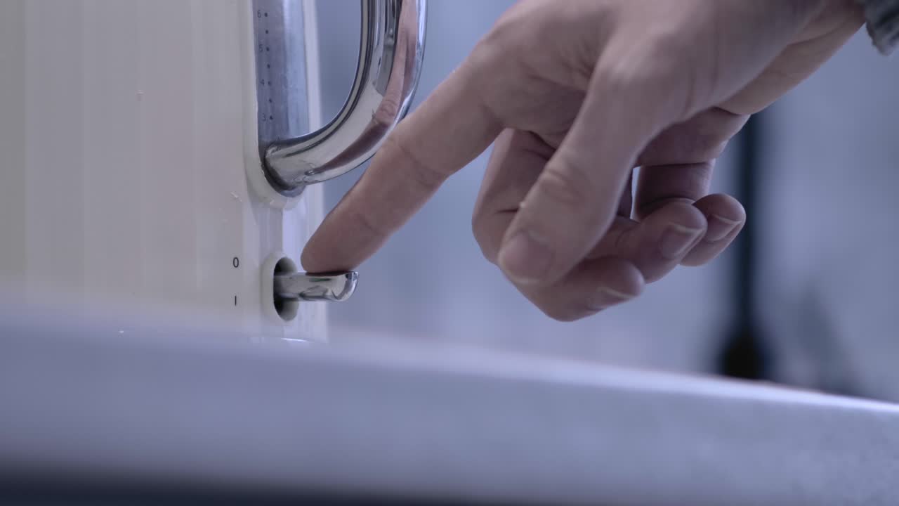 From low angle, a man's hand reaches into frame and turns on the switch of a kettle which lights up red.