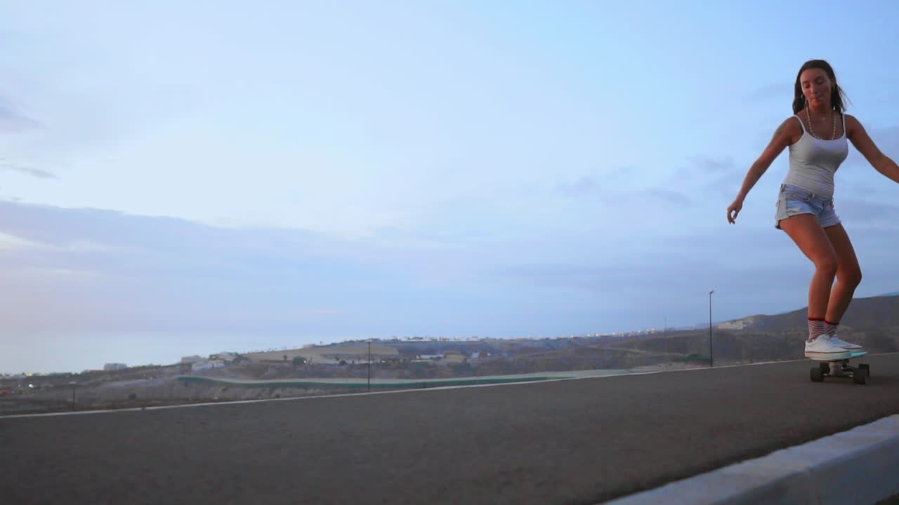 Riding her skateboard gracefully in shorts at sunset, a stunning young and fashionable skateboarder glides along a mountain road, capturing the mountains' majestic view in slow motion