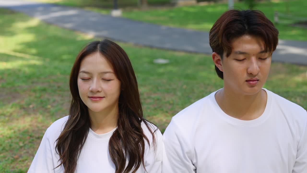 คู่รักชาวเอเชียอายุน้อยนั่งสมาธิโดยหลับตาในสวนที่มีแดดส่องภายใต้แสงจ้า