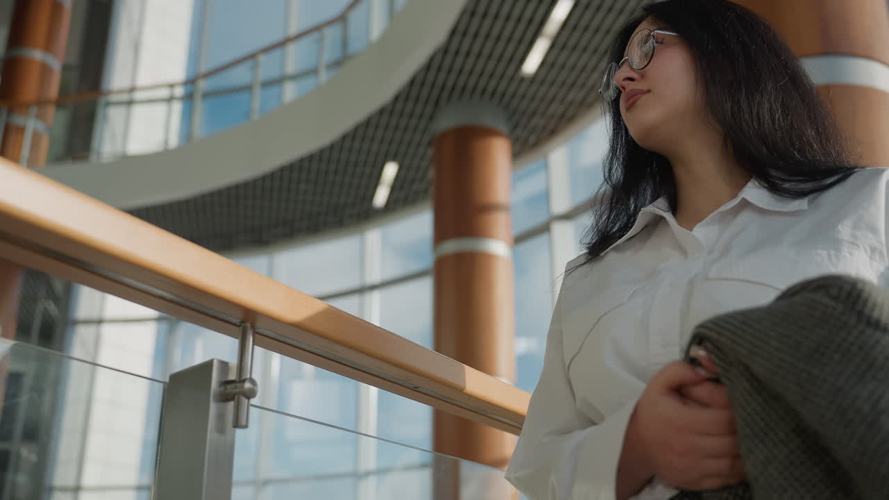 Young woman in white shirt and eyeglasses walking indoors close to glass railing with metal and wood elements, carrying checkered coat in arm while glancing upward in modern spacious mall