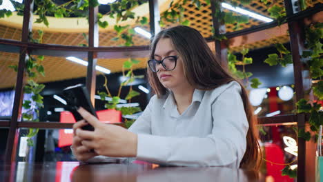 young adult seated in decorative wooden sit-out, operating phone with a subtle smile, in a well-decorated restaurant within a bustling mall, with a vibrant, lively atmosphere in the background