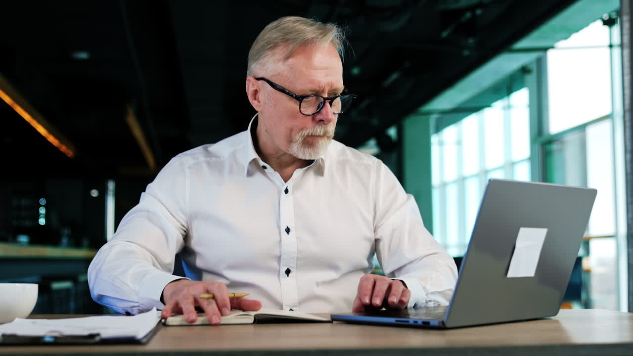 Focused male entrepreneur sitting at desk taking notes into his paper notebook. Man checks the information in laptop and writes down in a book.