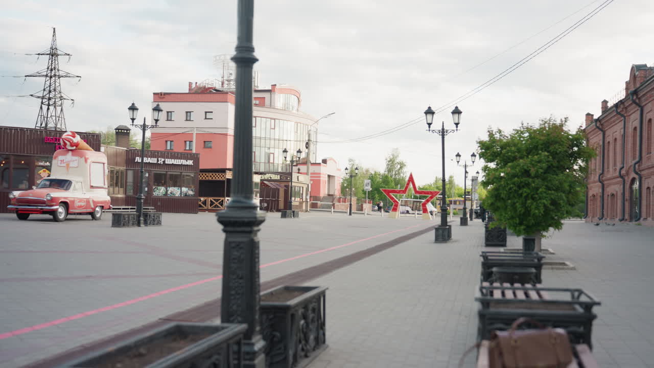 Камера снимает улицу в противоположном направлении: маленькое такси везет длинное мороженое-эскимо, проезжая мимо современного красного здания, в то время как женщина пользуется телефоном, а мужчина идет поблизости под мягким облачным дневным светом