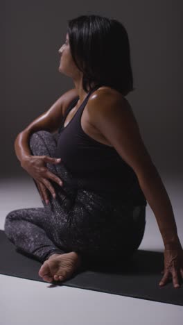 vertical video studio shot of mature woman wearing gym fitness clothing sitting on floor doing pilates stretching exercises