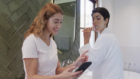 Feliz-Pareja-De-Lesbianas-Caucásicas-Cepillándose-Los-Dientes-Juntas-Y-Usando-Un-Teléfono-Inteligente-En-Un-Baño-Soleado