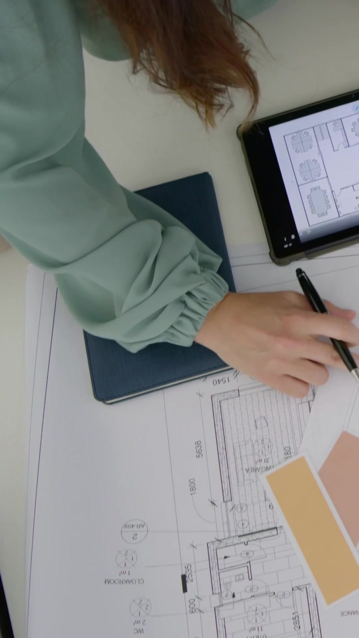 Vertical video: Holding pen on plan, woman in green top marking at desk, checking swatch for color