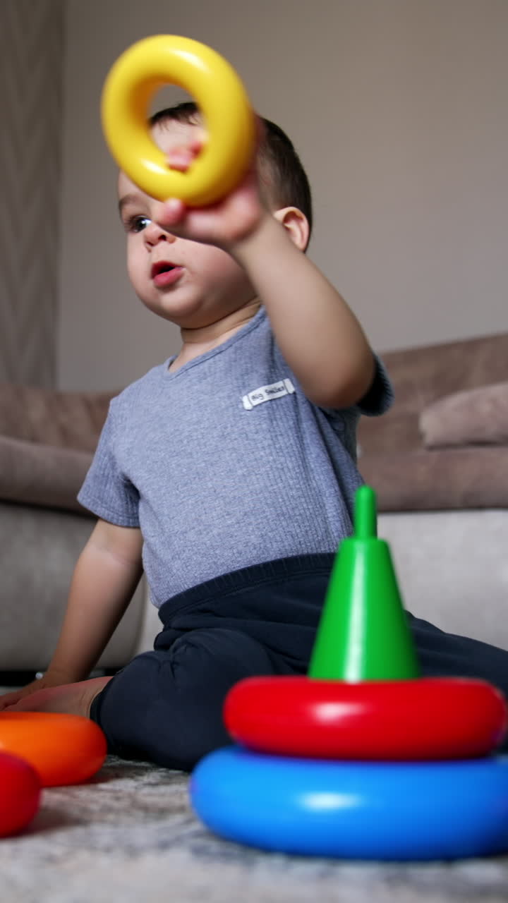 Calm serious baby boy plays on the floor. Kid constructs toy pyramids stacking the rings. Black cat sits beside watching the child. Low angle view. Vertical video
