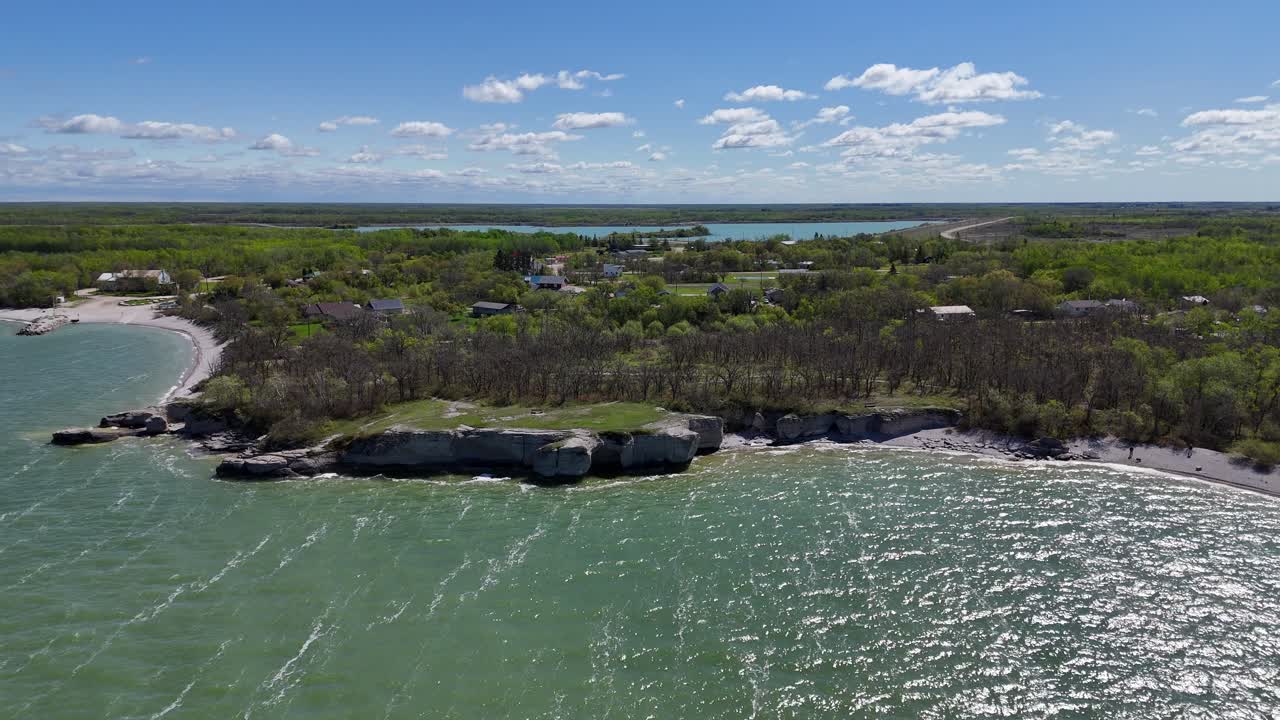 Ripresa drone più lontana dalle scogliere calcaree a Steep Rock Manitoba, orbitando a sinistra, che mostra l'acqua verde del Lago Manitoba, la costa e gli alberi in una soleggiata giornata estiva.