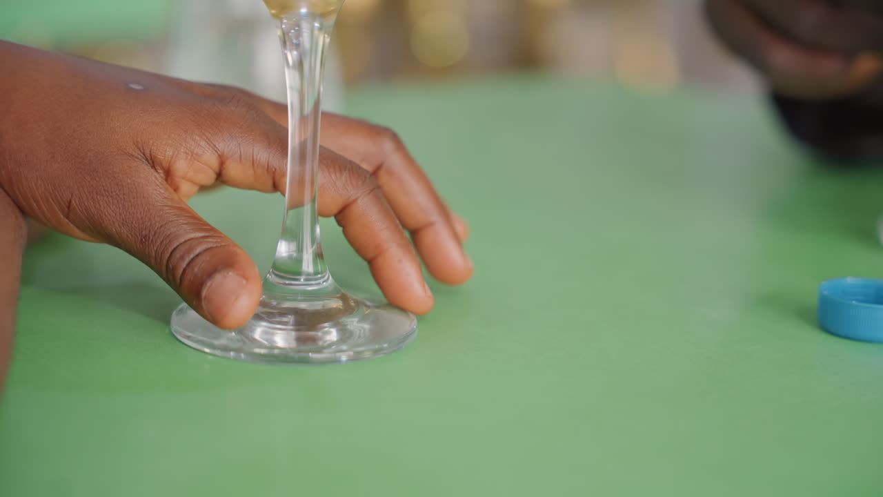 Black couple hands touch wine glass green, playful exchange near blue bottle cap on green table, two sets of fingers interacting in candid cafe scene, flirtatious gesture and shared moment, cozy