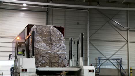 Forklift-loading-a-large-wrapped-pallet-into-a-truck-at-an-indoor-warehouse,-worker-visible