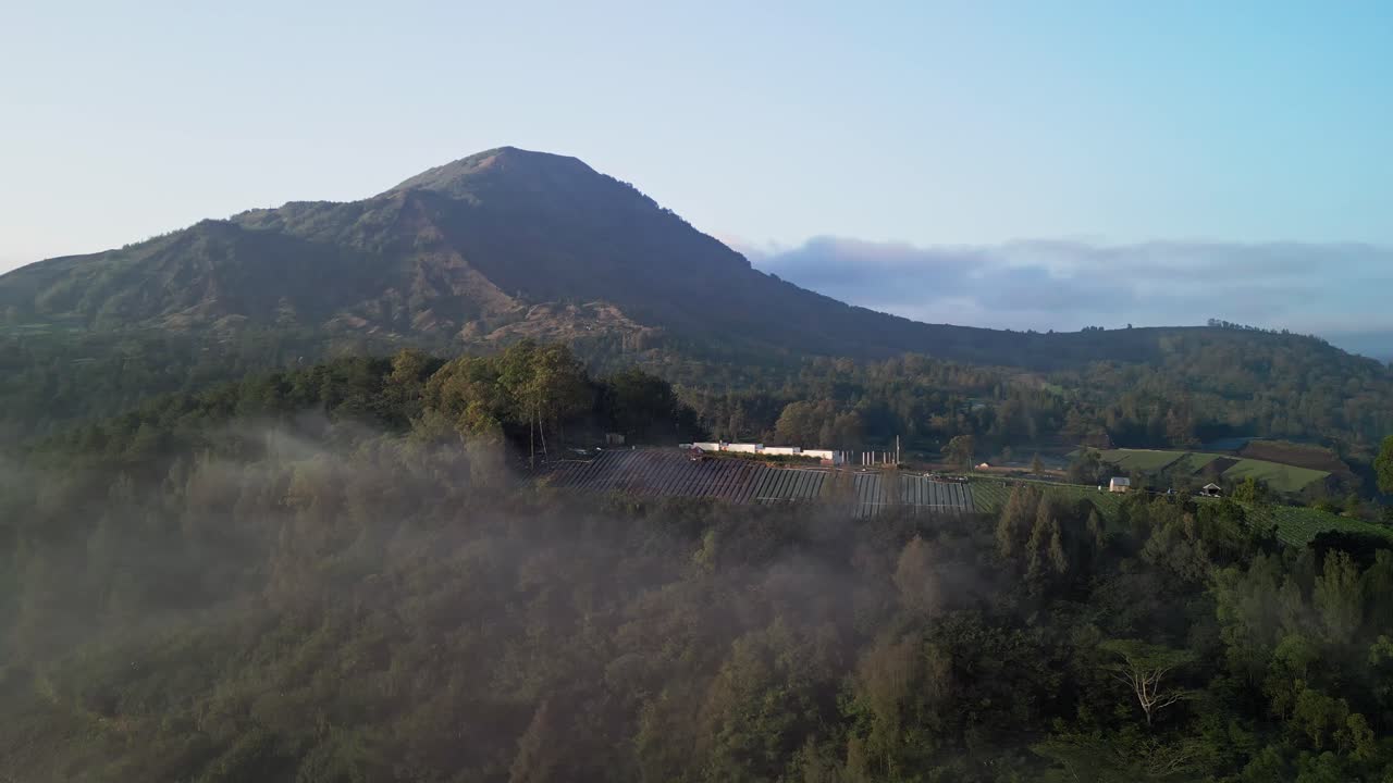Early morning aerial view of Mount Batur showing forested hills, small farming fields and gentle low clouds, creating a peaceful natural landscape around the volcano slopes in Bali Indonesia