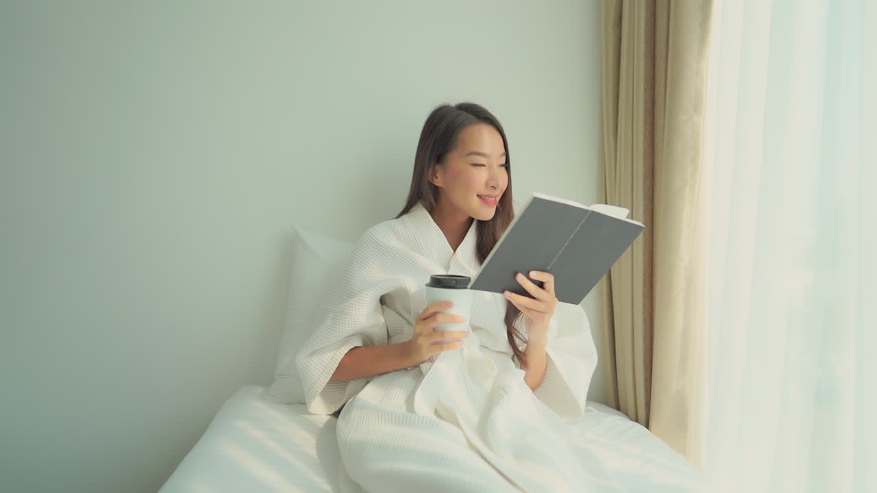 Attractive Asian lady sits on bed, drinks coffee and reads book in room