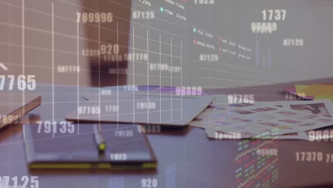 Animation-of-numbers,-graphs,-trading-boards-over-laptop-dairy,-paper-and-photographs-on-desk