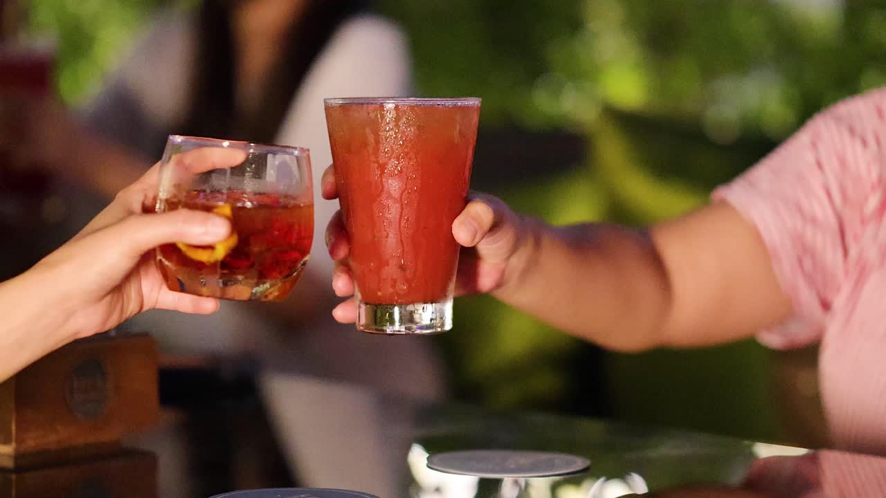 Two people clink cocktail glasses at an outdoor bar, evening light, relaxed social atmosphere