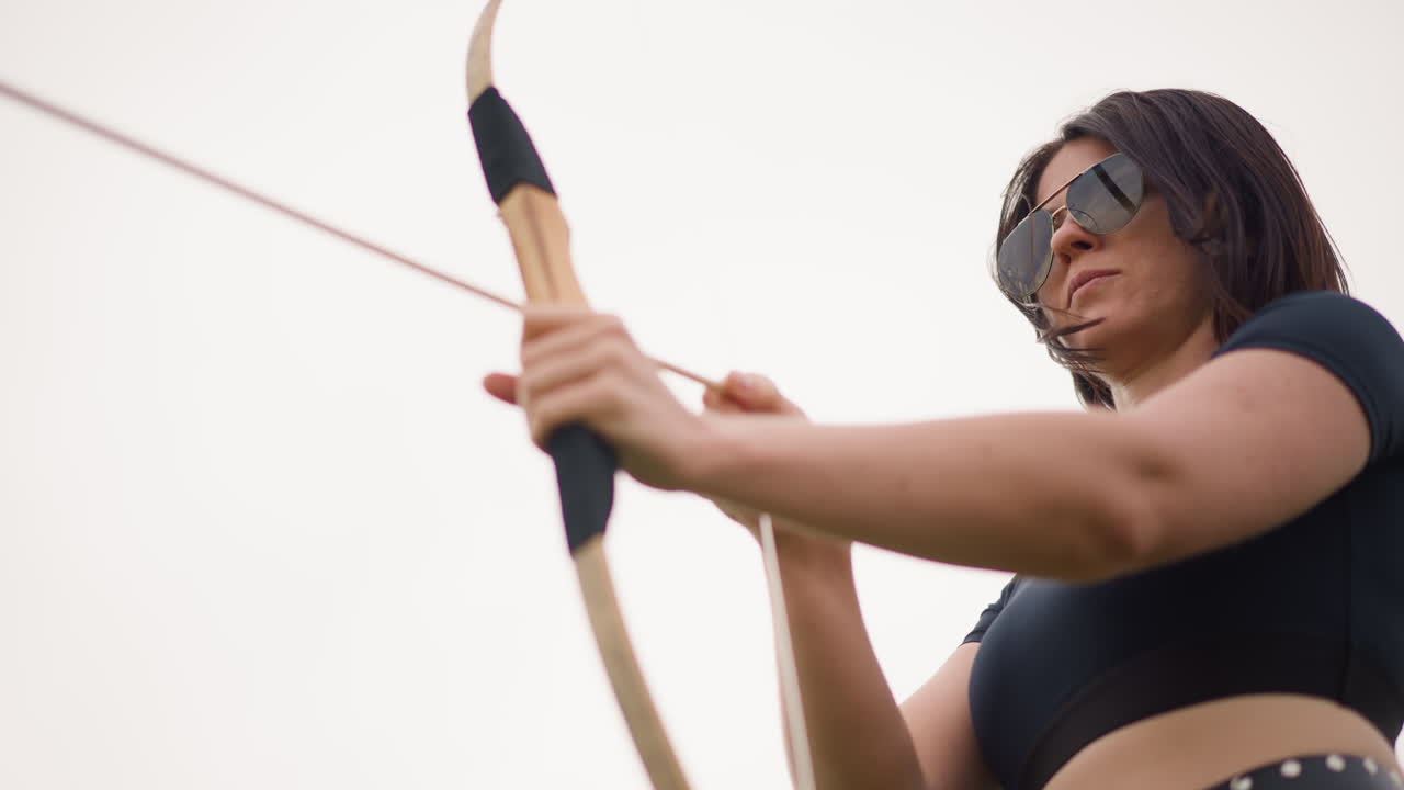 Donna che mira con un arco di legno, pronta a scoccare la freccia, con occhiali da sole e mascella serrata. Scatto dal basso che cattura la tensione della trazione e la freccia in posizione di messa a fuoco competitiva, tensione muscolare nell'avambraccio, sfondo di campo aperto